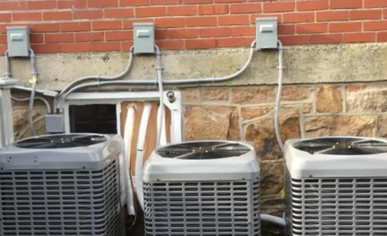 Three outdoor air conditioning units are lined up in front of a brick building.